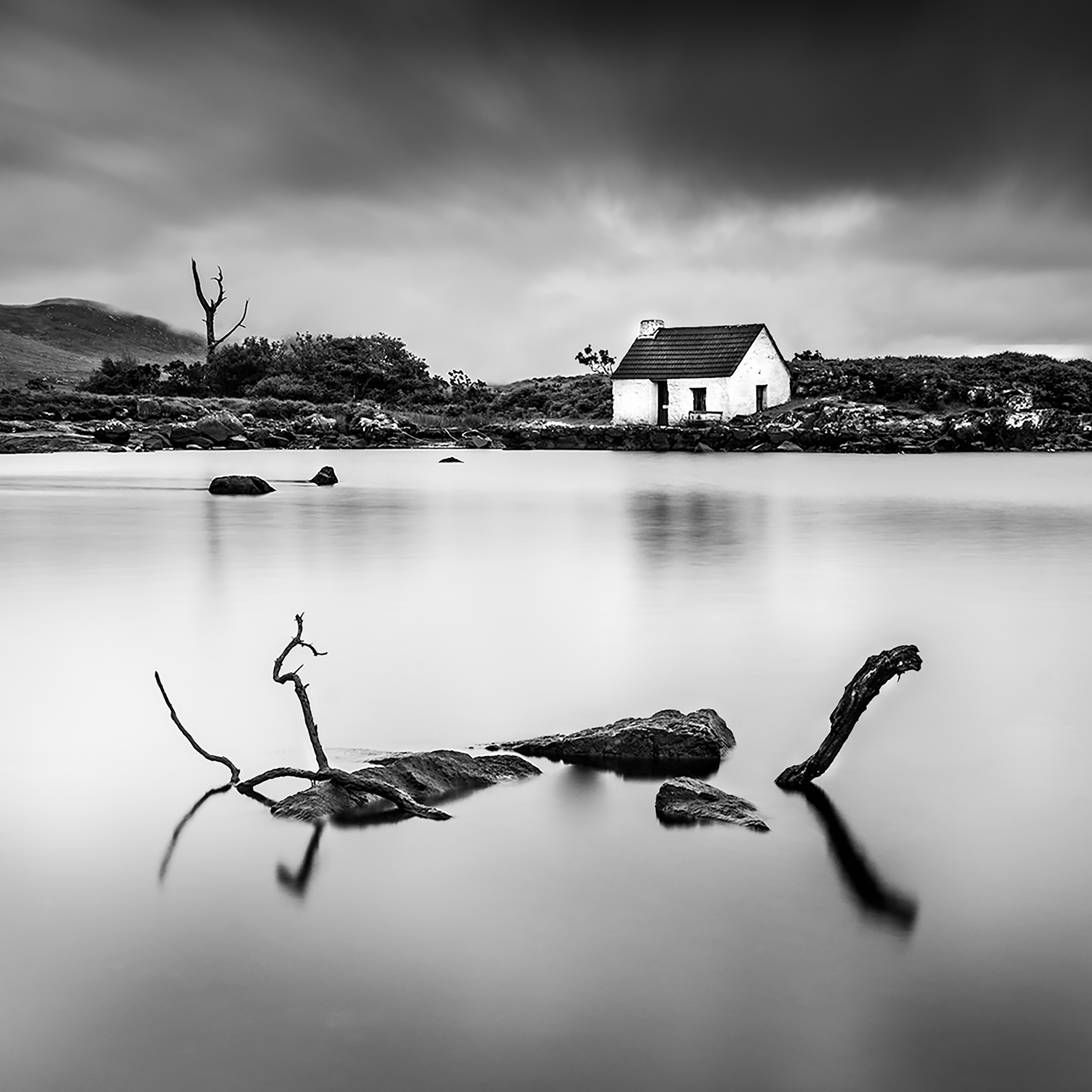 Connemara Fishing Cottage — a white cottage across still water with branches and rocks in the foreground